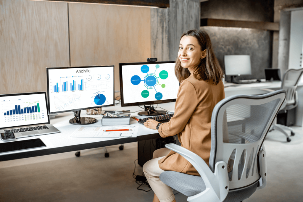 Een cybersecurity professional werkt achter haar bureau met twee beeldschermen waarop dashboards te zien zijn. Ze kijkt in de camera, wat vertrouwen en expertise uitstraalt.