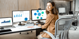 Een cybersecurity professional werkt achter haar bureau met twee beeldschermen waarop dashboards te zien zijn. Ze kijkt in de camera, wat vertrouwen en expertise uitstraalt.